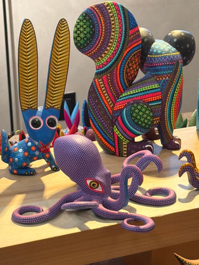 This image showcases a vibrant display of intricately carved and painted wooden folk art, known as alebrijes, against a light-colored background, likely a wall or shelf. The photo is oriented vertically, with the sculptures appearing to be displayed on a horizontal surface.
The most prominent sculpture in the upper left is a large, stylized animal, possibly an armadillo or similar creature, with a rounded body and stubby limbs. It is adorned with incredibly detailed, repetitive geometric patterns in a riot of bright colors including purples, blues, greens, oranges, reds, and yellows. Its head, partially visible at the top, is dark with white dot patterns.
To the right, a purple octopus sculpture captures attention with its complex swirling tentacles and a single, large, striking eye featuring a yellow iris and a red pupil. The entire surface of the octopus is covered in a uniform pattern of small white dots, giving it texture.
In the lower part of the image, a blue rabbit-like creature is visible. It has large, elongated golden ears with carved details resembling feathers or leaves. Its face features prominent, wide-set black eyes with white reflections. Its body is adorned with white and orange dot patterns. Other smaller, colorful figures are partially visible in the background, adding to the bustling collection.
The objects are brightly lit, casting distinct shadows on the display surface, suggesting a strong light source, possibly from the right. There is no visible text in the image. The style and vibrant detail are characteristic of alebrijes from San Martín Tilcajete, Mexico.