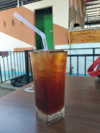 The image features a glass of iced tea with a white straw, placed on a wooden table. The drink appears to be mostly consumed, with ice visible at the top. The background includes a partially visible building with brown and white exterior paint and a green interior visible through a doorway. A black metal railing is also visible, suggesting an outdoor or semi-outdoor setting. Given the city mentioned, Matang, Indonesia, it is highly possible that the image depicts a local establishment.