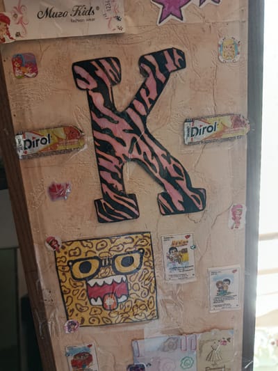 The image shows a close-up, vertical, eye-level view of a light-brown, textured surface, likely a wall or board, decorated with various stickers, papers, and a large letter "K". The lighting suggests it is daytime, with sunlight casting shadows and highlights, particularly on the right side of the frame.
Dominating the center is a large, uppercase letter "K" with a pink and black tiger-striped pattern, covered in clear plastic. To the left of the "K" are two small stickers: one with a pink border and an image of two cartoon girls with red hair, and another featuring a red plastic heart shape with some glitter.
Above the "K" is a white paper tag with "Muzo Kids" printed in a stylized font, along with "fashion wear" below it. There are also small decorative elements on this tag, including a cartoon figure and some floral designs.
To the right of the "K" is a small, circular sticker of a blonde cartoon girl with a blue ribbon in her hair. Below this, and slightly to the right of the "K," is a small, rectangular wrapper for "Dirol" gum, featuring images of fruit.
Further down and to the left of the "K" is a drawing of a cartoon face with leopard-print patterned skin, large yellow-rimmed glasses with black pupils, and a wide, open mouth revealing red lips and a white tongue. This drawing is also covered in clear plastic.
Scattered around the drawing and the "K" are several smaller stickers, including cartoon characters and small printed cards with text in Russian and English, some featuring couples or single figures. One card on the right has the text "Любовь это... Love is..." and a picture of a couple embracing.
The bottom of the image reveals parts of other decorative items, including a drawing of a dress and the word "Dominique."
The scene is indoors, with the light suggesting a window is present to the right. The overall aesthetic is childlike and decorative, suggesting a personalized space. There are no visible indications of the city of Dachne, Ukraine within the image itself.