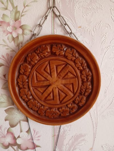 A carved wooden disc with a sun symbol is hanging on a wall with floral wallpaper. The disc is circular with a raised rim and intricate carvings of leaves around the edge. The central carving is a stylized sun with eight rays, each composed of three triangular segments. The disc is suspended by a metal chain. The wallpaper has a pale pink background with a repeating pattern of stylized flowers and leaves in muted pink, green, and white. The overall impression is that of a decorative item displayed in a domestic setting. The image was taken in Kyshtym, Russia.
