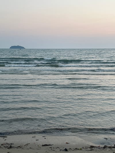 The image shows a view of the ocean and beach in Baan Klong Prao, Thailand. The beach is on the right side, transitioning to the ocean on the left. Gentle waves are visible throughout the water. The sky is pale, with a possible sunset or sunrise occurring. There is a landmass in the distance, partially obscured by the sky's haze. Debris litters the shoreline.