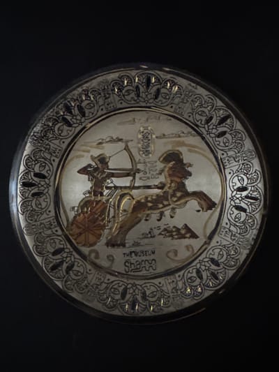 The image shows a decorative plate with an Egyptian-themed design. The plate is circular and has a metallic sheen, possibly silver or pewter. The central motif depicts an ancient Egyptian chariot with a warrior aiming a bow and arrow, pulled by a horse. Hieroglyphics are visible above the chariot and in the decorative border of the plate. The border is intricate, featuring stylized patterns and what appear to be lotus flowers. Text on the lower part of the plate reads "THE MUSEUM" and "Sharps". The plate is set against a plain black background. There is no indication of its location in Grodzisk Wielkopolski, Poland.