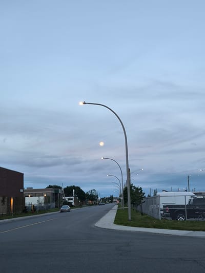The image shows a street scene in Dorval, Canada, during twilight. A road stretches into the distance, flanked by sidewalks, grass, and buildings. Several streetlights are visible, some illuminated. A silver sedan is parked on the right side of the road, and further down, a white van and a black pickup truck are parked behind a chain-link fence. The sky is overcast with a soft glow from the setting sun or streetlights, and a faint, pale moon is visible. Trees line the street, and a brick building is prominent on the left side of the image.