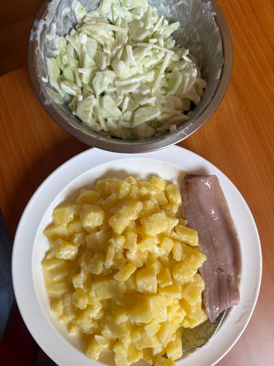 This media file shows a top-down view of a meal on a wooden table. The meal consists of a metal bowl filled with a light-colored salad, likely a coleslaw or cucumber salad with a creamy dressing, and a white plate with a piece of fish and a mound of cubed potatoes. The fish appears to be a fillet, possibly cured or smoked, with a pinkish-brown hue. The potatoes are cooked and appear to be dressed with a creamy sauce, suggesting they might be part of a potato salad. A spoon is partially visible on the plate, resting amongst the potatoes. The lighting suggests it is daytime. The setting is casual, likely a home or a simple restaurant. The focus is entirely on the food, with no people visible. The colors are natural, with the warm tones of the wooden table contrasting with the lighter colors of the food. There is no visible text in the image.