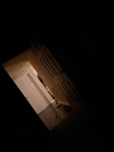The image shows an interior view of a hallway with two doors, one white and one brown, set at an angle. The scene is dimly lit, with the majority of the frame consumed by darkness. A string of small lights is visible along the top of the white door, suggesting a festive or decorative element. The white door is a standard interior door with paneling and a silver doorknob. The brown door, to the right, also has paneling and appears to be ajar. The flooring is a dark color, possibly hardwood or laminate. The background is obscured by shadow, but hints of a room beyond the brown door suggest it leads to another space. The image is angled, giving a sense of looking into the scene from above or an unusual perspective. The location is identified as Sandy Springs, United States of America.