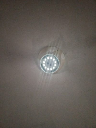 A white, circular surface-mounted LED light fixture is centrally located on a light-colored, uniform ceiling in Keo Kouang, Laos. The fixture contains approximately twelve to fourteen illuminated individual white LEDs arranged in a concentric circular pattern, emitting distinct light rays downwards. The ceiling appears smooth and devoid of other objects. No individuals or additional subjects are present in the image, which is captured from a low-angle perspective looking directly upwards at the active light source.