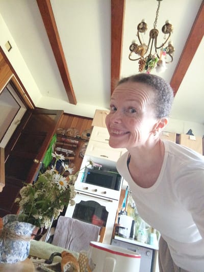 The image captures a selfie taken by a woman indoors, likely in a residential setting. She is positioned in the lower right of the frame, looking towards the camera with a wide smile. The woman has short, graying hair and is wearing a white long-sleeved shirt. Her left ear is visible, adorned with a small earring.

The background suggests a kitchen or dining area. To the left of the woman, a bouquet of white daisies and other greenery is placed in a blue and white patterned pot, tied with twine. Behind the flowers, a dark wooden door is visible.

In the middle ground, a white kitchen appliance with a black microwave on top is present. Various items are displayed on shelves above and to the left of this appliance, including what appear to be plates or bowls and jars. A striped dishtowel is folded on a surface to the right of the flower arrangement.

The ceiling is white with exposed wooden beams, and a chandelier hangs from the ceiling. The overall environment appears to be well-lit, with natural light entering from an unseen source, creating highlights and shadows. The image does not provide any specific clues to indicate the location as Alexandrovskaya, Russia.