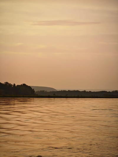 The image depicts a serene river scene at dusk. The sky is a soft, muted orange, with wispy clouds. A dark, dense line of trees forms the horizon, with rolling hills visible in the distance. The river in the foreground is calm, with gentle ripples reflecting the warm tones of the sky. No people or specific structures are visible, suggesting a natural, undeveloped landscape. The overall atmosphere is tranquil and picturesque, characteristic of the natural beauty found in and around Maloe Nakhval'skoe, Russia, during sunset.