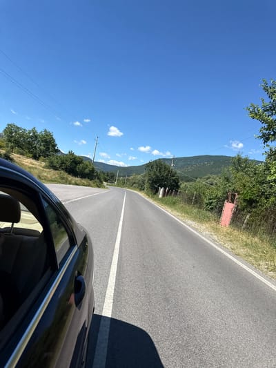 The image depicts a scenic view of a winding asphalt road with a white line down the center, curving through a lush green mountainous landscape under a clear blue sky with scattered white clouds. The perspective is from inside a dark-colored car, showing the side and rear-view window. The road passes by dry, grassy hillsides dotted with trees and bushes on the left, and a fence line with vegetation and a red metal gate on the right. In the distance, rolling green mountains rise against the horizon. Power lines are visible stretching across the sky. The scene appears to be a rural setting, likely in the vicinity of Sotirya, Bulgaria, given the natural beauty and the type of road. There are no people visible in the image.