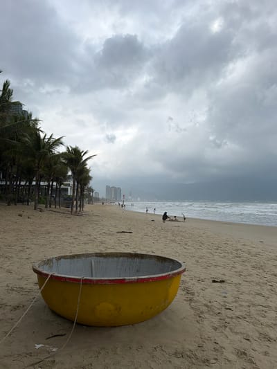 The image depicts a beach scene in An Hải Ward, Vietnam, under heavily overcast skies. In the immediate foreground, a circular, yellow-painted coracle (basket boat) with a grey interior and a red rim rests on the wet, light-tan sand. Two thin, light-colored ropes extend from the boat onto the sand. The sandy beach stretches into the midground towards the right, meeting the ocean with white-capped waves breaking on the shore.

Along the midground beachfront, a line of tall palm trees with dark green fronds runs parallel to the left, behind which multi-story buildings are partially visible. Several individuals are present on the beach: a figure in dark clothing is crouched near the waterline on the right, seemingly interacting with another smaller figure, possibly a child. Further to the left, multiple smaller figures are discernible walking or standing along the shoreline. In the distant background, a dense cluster of high-rise buildings forms a cityscape along the coastline, partially obscured by the atmospheric conditions. The ocean surface appears a muted grey-blue under the expansive, dark grey cloud cover.