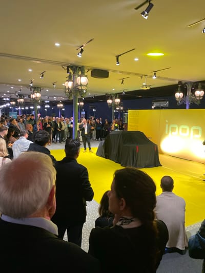 This image depicts an indoor event, likely a product launch or conference, taking place in Mulhouse, France. A large gathering of people, predominantly adults, are visible from behind, facing a brightly lit stage. The stage features a vibrant yellow backdrop with a large, stylized logo that appears to read "iqop" or a similar word, and possibly "EDF".

A prominent feature on the stage is a large object covered with a black sheet, hinting at an imminent unveiling. Adjacent to this covered object, a banner is visible with key details of the event. The text on the banner clearly states "PREMIERE MONDIALE" (World Premiere), "23 AVRIL 2023" (April 23, 2023), "L'AUTOMOBILE" (The Automobile), and "MULHOUSE", indicating that this is the global debut of a new car in Mulhouse on that date.

The venue is an event hall with decorative lighting fixtures, including modern spotlights on the ceiling and ornate, street-lamp-style luminaires along the walls, providing ample illumination. The overall setting suggests a formal and anticipatory atmosphere. The crowd appears attentive, focused on the stage where the presentation is taking place.
