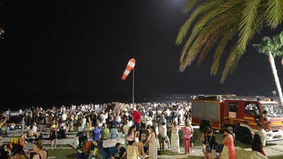The image captures a nighttime scene on a beach in Almuñécar, Spain, likely during a festival or public gathering. A large crowd of people is visible, many seated or standing on the sand and a low concrete barrier. Palm trees, including a large one in the foreground to the right, frame the scene. A prominent object is a red fire truck with "BOMBEROS" and "ALMUÑÉCAR" written on its side, along with a "112" emergency number emblem. A red and white striped windsock is also visible on a pole. The ocean and a dark sky are in the background. People are dressed in casual clothing, and some are interacting in small groups. The overall atmosphere appears to be lively, with the presence of the fire truck suggesting a possible organized event or the need for emergency services.
