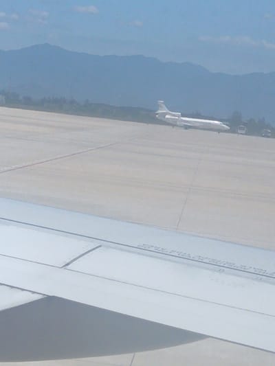 The image shows an airplane on a tarmac with mountains in the background. The airplane is white with a blue stripe. There are also two buses visible on the tarmac. The sky is blue with some clouds. The image appears to be taken from an airplane window, as a portion of an airplane wing is visible in the foreground. The scene is likely at an airport in Bắc Cam Ranh, Vietnam, given the presence of mountains that are characteristic of the region. The lighting suggests it is daytime. The focus is on the airplane and the surrounding airport environment.