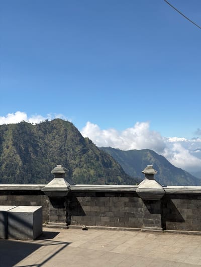 The image depicts a scenic view from a walled overlook in Cemorolawang, Indonesia. In the foreground, a dark stone wall with decorative pillars forms a barrier. To the left, two large, grey, concrete cube-shaped structures are visible, casting long shadows. The ground is paved with square tiles. Beyond the wall, a vast mountain range dominates the background. The mountains are covered in dense green vegetation, with some areas appearing rocky and barren. White clouds are scattered across a clear blue sky, with a thin wire or cable visible in the upper right corner. The overall scene suggests a viewpoint offering a panoramic vista of the natural landscape surrounding the town.