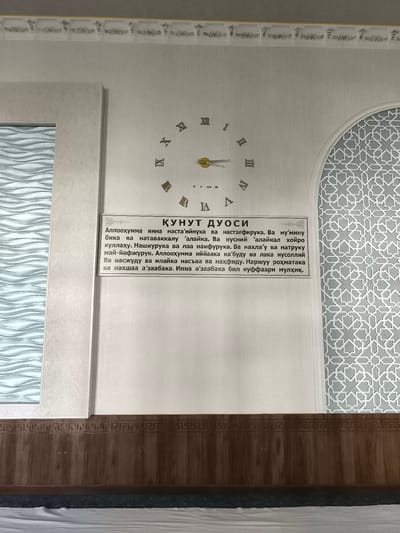 The image shows a close-up of a wall in a building in Бабаходжа, Uzbekistan. The wall is decorated with a large, modern Roman numeral clock and a framed text in an unfamiliar script. The clock's hands are gold and point to approximately 10:28. To the right of the clock, there is an arched alcove with a repeating geometric pattern in white and gray. Below the clock and text is a dark brown wooden wainscoting with a decorative border. The overall environment appears to be indoors, possibly a religious or community building, given the decorative elements.