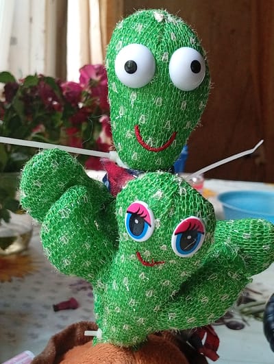 Two plush toy cacti with large googly eyes and smiling mouths are positioned in the foreground. The cactus in the front has blue and pink eyes, while the one behind it has black and white eyes. Both cacti are green and textured with white sparkly threads. They appear to be part of a display or decoration. In the background, a blurred arrangement of pink flowers in a pot is visible, along with curtains and a wooden surface. A blue bowl and a table with a patterned cloth are also present. The overall scene suggests an indoor setting, possibly a home or a shop in Berezivka, Ukraine.