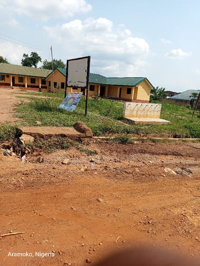The scene is a dirt road in front of a building complex in Ilobu, Nigeria. The buildings are a series of low, rectangular structures with green roofs, likely classrooms. A white sign with black lettering is mounted on a post in front of the buildings. A poster on the sign is partially visible. There are small trees and shrubs around the buildings. The road is covered in dirt and debris. There are no people visible in the scene. The sky is partially cloudy. The coordinates are 7.835 latitude and 4.481 longitude.