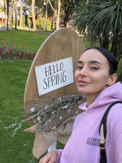 A woman wearing a light purple hoodie and a black shoulder strap poses for a selfie next to a table with a "Hello Spring" sign on it. The table is decorated with a burlap bow and olive branches. The scene is set in a park in Grad Opatija, Croatia, recognizable by the well-maintained lawn, flower beds, trees, and buildings in the background.