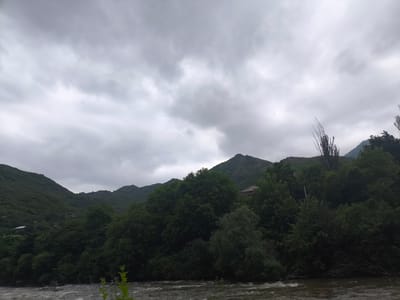 The image depicts a wide, turbulent river flowing through a valley. Lush green trees and dense foliage line the riverbanks, climbing up the sides of rolling, verdant mountains in the background. The sky is overcast with thick, grey clouds, suggesting a dim or possibly rainy day. In the mid-ground, peeking through the trees on the right side, is a hint of a building with a reddish-brown roof, suggesting a settlement or habitation within this natural landscape. The scene is consistent with the environment found in and around the city of Tumanyan, Armenia, known for its mountainous terrain and the Debed River.