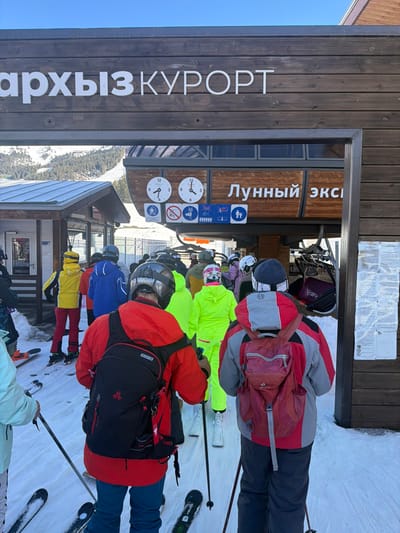 The image depicts a queue of approximately 15-20 individuals in ski gear, waiting to board a ski lift at the Arkhyz Resort, specifically at the "Lunny Ekspress" (Lunar Express) lift station. This location is situated within the mountainous region associated with Lunnaya Polyana, Russia.
The scene is viewed from behind two skiers in the immediate foreground, both wearing helmets and backpacks, holding ski poles, and standing on skis. The skier on the left wears a red jacket and blue pants with a black backpack. The skier on the right wears a grey and red jacket with a small red backpack.
Behind them, a line of skiers, also in helmets and varying bright ski apparel (including neon green, blue, yellow, and red jackets), are positioned on a snow-covered incline leading towards the lift entrance. Their skis are on the snow.
The ski lift station is a substantial wooden structure. Above the entrance, a sign partially visible reads "АРХЫЗ КУРОРТ" (Arkhyz Resort) on a horizontal wooden beam. Below this, on the station building, the words "ЛУННЫЙ ЭКС" (Lunny Ekspress) are visible in white lettering. Two circular clocks, displaying different times (approximately 9:00 and 1:00), are mounted on the wooden facade. Below the clocks are several pictographic safety signs, indicating rules such as "no standing," "no heavy luggage," "children must be accompanied," and "seated."
To the left of the main lift station, a smaller building with a sloped, snow-covered roof is partially visible. The overall environment is a bright, clear day, with snow covering the ground and distant mountains visible under a blue sky, characteristic of a winter ski resort.