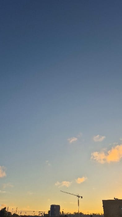A vast, gradient-filled sky dominates the scene, shifting from deep blue to pale yellow with hints of orange. Wispy clouds are scattered across the lighter section of the sky. The silhouette of a cityscape, including a crane, stands on the right edge, contrasting against the bright backdrop. Buildings and other structures are distinguishable.
