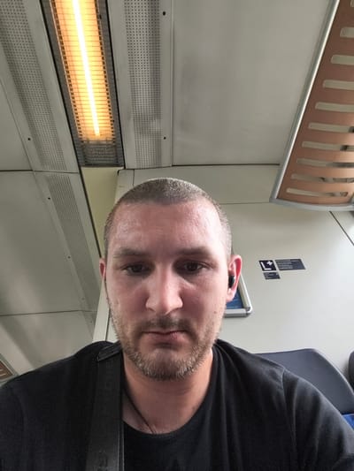 A man with a shaved head and stubble is pictured from the chest up, looking at the camera. He is wearing a black t-shirt and a black earbud is in his right ear. The background appears to be the interior of a train carriage, with a bright light fixture on the ceiling and the back of a seat visible. A sign with an exclamation mark and text is on the wall to the right of the man. The image was taken in Züssow, Germany.