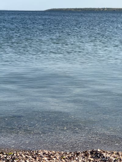 The image shows a body of water with a shoreline in the foreground and a distant landmass in the background. The water is calm with gentle ripples, reflecting the light blue sky. The shoreline is composed of small pebbles and shells, with some patches of greenery. The distant landmass appears to be a low-lying hill or bluff covered in vegetation. The scene is set in Ukrainka, Ukraine, likely a coastal or riverside area. There are no people or specific objects of interest visible in the image.