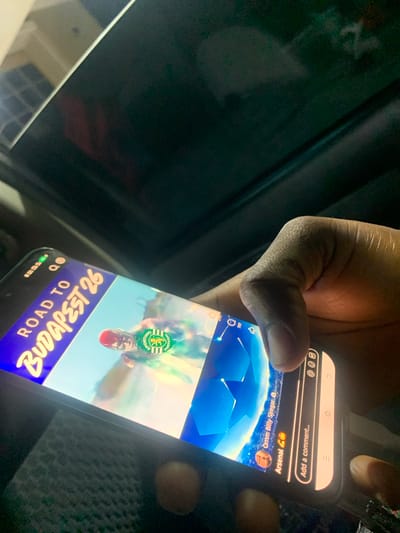 A close-up shows a hand holding a smartphone. The phone's screen displays a social media post. The post features a graphic with the text "ROAD TO BUDAPEST 26," a person wearing a green and white Sporting CP jersey, and the Sporting CP logo. Above the graphic, the social media interface shows the user "Omm Billy Siregar" and "Arsenal," suggesting a football connection. The "Add a comment..." field indicates the user is viewing or about to interact with the post. The background is dark and unclear. The location in the city of Jiru, Nigeria, is not determinable from the information in the picture.