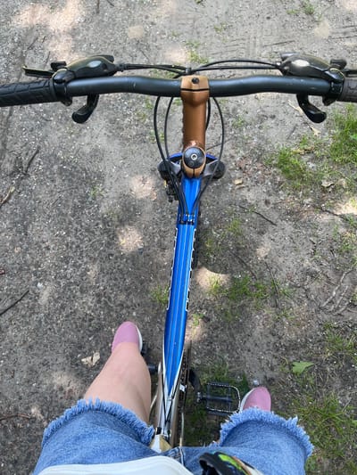 The image shows a first-person perspective of someone riding a bicycle on a dirt path. The handlebars, front of the bicycle frame, and the rider's legs are visible. The rider is wearing denim shorts with a frayed hem and pink sneakers. The path appears to be in an outdoor, possibly wooded area, with grass and dirt. The location is identified as Pohreby, Ukraine.