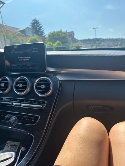 The image shows the interior of a Mercedes-Benz C-Class car on a sunny day, with the location identified as Räterschen, Switzerland. The car's dashboard is visible, featuring a central infotainment screen displaying "Bluetooth Audio-Gerät wurde angehalten." Below the screen are climate control vents and buttons. The digital clock on the dashboard reads 15:38. The view outside the car through the windshield shows a suburban area with houses and trees under a clear blue sky. The legs of a person are visible in the foreground, suggesting the photographer is a passenger. The overall scene is static and focused on the car's interior and immediate surroundings.