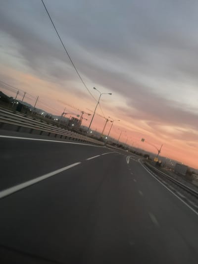 The image depicts a perspective from a moving vehicle on a highway, likely during dusk or dawn, given the soft, warm hues of orange and pink in the sky against a backdrop of gray clouds. The highway itself is asphalt, with white lane markers visible. To the left of the road, there's a barrier, beyond which a cityscape or construction site is visible. Several streetlights are illuminated, and construction cranes stand tall against the colorful sky, suggesting ongoing development. Power lines and poles are also present, adding to the urban or semi-urban environment. The overall impression is one of a journey or transit through an evolving landscape as day transitions into night or vice versa. The tilted angle of the photograph, combined with the motion blur, enhances the sense of movement and dynamism. There is no visible text.
