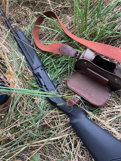 A black semi-automatic Beretta shotgun lies on the ground, oriented diagonally with the barrel pointing towards the upper left. Its action is open, exposing the bolt and chamber area. Adjacent to the shotgun, to the right, is an open, olive-green canvas and brown leather cartridge bag, with its top flap folded back to reveal an internal compartment. An orange-brown woven fabric strap with brown leather attachments, consistent with a gun sling or bag strap, is draped over the grass behind the shotgun and bag. The items are situated on an uneven surface of tall, mixed dry and green grasses and other low-lying vegetation. The scene depicts an outdoor, rural environment, consistent with fields or natural areas found in the vicinity of Skipsea, United Kingdom. No individuals are present, and all objects are static.