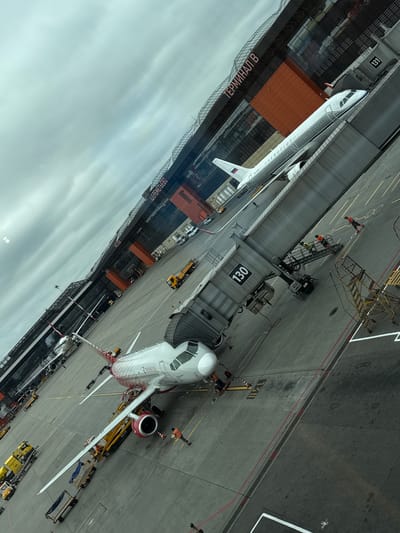 The image shows an airport scene at Sheremetyevo International Airport (SVO) in Khimki, Russia, near Rzhavki 2. Two airplanes are visible on the tarmac. One airplane, identified by "AEROFLOT" on its fuselage and "В-2710" on its tail, is connected to a jet bridge labeled "131". Another airplane, with red and white livery and the text "ROSSIYA" on its side, is also parked and appears to be undergoing servicing, with workers and ground equipment around it. Several service vehicles, including yellow trucks and a luggage cart, are present on the tarmac. The airport terminal building, marked with "ТЕРМИНАЛ В" and "МОСКВА", is visible in the background under a cloudy sky. Several airport workers in high-visibility vests are engaged in various tasks on the ground and on the jet bridge. The overall environment is an active airport operational area.