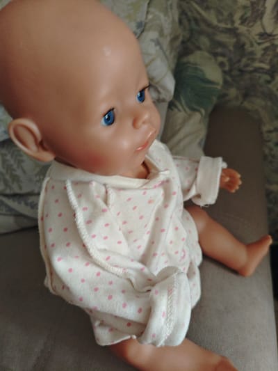 A baby doll with blue eyes and a bald head wears a white outfit with pink polka dots. The doll is lying on its side on a gray surface, with its right arm extended. In the background, a patterned fabric is visible. The image appears to be taken indoors. There is no information available about the location in the city of Berdsk municipality, Russia.