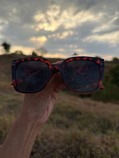 A hand holds a pair of tortoiseshell sunglasses with dark lenses in the foreground. The sunglasses are the primary focus of the image, with their reflective lenses showing a distorted image of the surroundings. In the background, the scene depicts a cloudy sky with patches of bright sunlight, suggesting it is either sunrise or sunset. The sky is a mix of grays and golden hues. To the right of the sunglasses, a blurred background of dry, brown vegetation and greenery can be observed, indicating an outdoor, possibly rural or natural setting. The lighting and the cloudy sky suggest a warm, possibly humid climate typical of Indonesia, specifically Kubutambahan. There are no visible text elements in the image. The overall impression is one of a casual moment, perhaps enjoying the outdoors during a picturesque time of day.