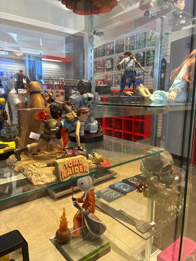 In an indoor display case, a variety of collectible figurines and merchandise are showcased. The primary focus is on a "Tomb Raider" themed display featuring a Lara Croft figurine in an action pose, an artifact, and the "Tomb Raider" logo. Adjacent to this, a stylized skull figurine with moss-like accents is positioned on a shelf. To the left, a figure resembling Sam from the movie "Trick 'r Treat" is depicted with a campfire accessory. Further back, other figures and media, possibly video games or movies, are visible on shelves, creating a densely populated retail display.