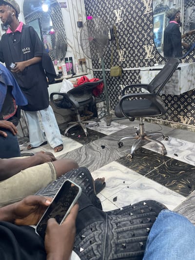 The image depicts an indoor scene inside what appears to be a barber shop in Safari, Nigeria. The shop features a distinctive black and white patterned wallpaper with a repeating "LV" monogram design. Several individuals are present. In the background, a barber, wearing a black smock with "OLA'S HAIR CUT" printed on it and a pink collar, is holding a hair clipper and appears to be working. A barber chair with a black mesh seat and a metal base with wheels is positioned in the center of the frame. A large oscillating fan is visible to the left. The floor is tiled with a pattern of black, white, and grey marbled squares. In the foreground, two individuals are seated, one holding a smartphone with a case displaying the same monogram pattern as the wallpaper. The overall environment suggests a functional and somewhat decorated space for hair services.