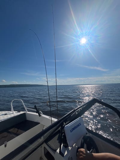 This media file depicts a daytime scene from a boat on a body of water, likely during a fishing trip. The sun is shining brightly, creating a strong lens flare across the upper left portion of the image and reflecting off the water's surface. The sky is a clear, bright blue with some scattered white clouds.
The main subjects are fishing rods positioned in rod holders on the boat. Two fishing rods are clearly visible, extending upwards and out over the water. One rod is bent, suggesting it might be in use or has a weight attached. A fishing reel is visible on one of the rods.
The boat itself is partially shown, with the console and part of the seating area visible on the right side of the frame. A person's hand is seen on the steering wheel, indicating they are piloting the boat. A "LOWRANCE" branded fishfinder or GPS unit is prominently displayed on the console. The water is calm with ripples, reflecting the sunlight. In the distance, a hazy landmass or shoreline is visible along the horizon.
The overall mood is one of leisure and outdoor activity, specifically fishing. The bright sun suggests it is midday. The presence of fishing equipment and the boat indicates a recreational activity taking place on a lake, river, or sea. The visible text "LOWRANCE" is a brand name for marine electronics. Given the provided location context of Siukterka, Russia, this scene likely takes place on one of the local waterways in that region.