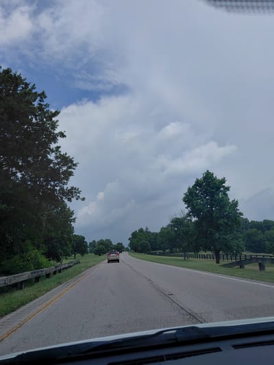 A vehicle drives down a two-lane asphalt road on a cloudy day in Paris, United States of America. The road is bordered by a guardrail on the left and trees and a black wooden fence on the right. In the distance, a few more vehicles are visible. The sky is filled with dramatic clouds, suggesting an approaching storm. The overall scene depicts a rural or semi-rural drive through a wooded area.