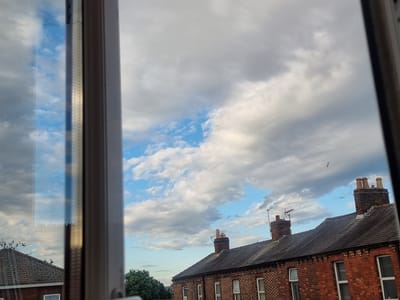 A view through a window shows a cloudy sky over a row of brick houses with dark roofs in Carlisle, United Kingdom. The sky is a mix of grey and white clouds with patches of blue visible. The houses have tiled roofs and brick facades. Several chimneys are visible on the rooftops, one with multiple stacks. A bird is in flight against the cloudy sky. To the left, a reflection in the window shows a portion of the sky and the window frame. The image appears to have been taken during daylight hours.