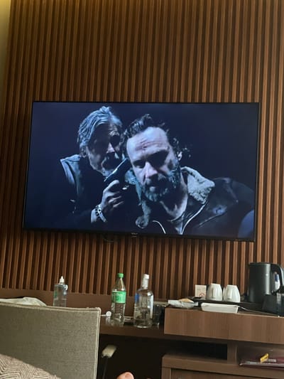 The image shows a television mounted on a horizontally ribbed wooden wall. The television screen displays a scene with two men, one older with gray hair and another younger man, both in what appears to be a dramatic scene. To the right of the television is a dark wooden cabinet. It holds items such as a black electric kettle, white cups, various packets, and bottles of liquid. A gray upholstered headboard of a bed and a person's hand are visible in the lower right corner of the frame. The image was taken indoors. There is no information to ascertain if this photo was taken in Otopeni, Romania.