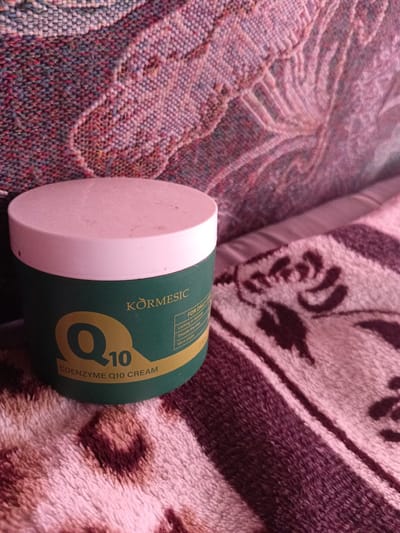 The image shows a jar of Kormesic Coenzyme Q10 Cream. The jar is green with a white lid. The text on the jar is in English and indicates that the cream is for daily care, locks in moisture, smooths fine lines, and makes skin more elastic. The jar is placed on a patterned fabric surface, which appears to be a blanket or a couch. The lighting suggests it is indoors, with some ambient light creating shadows. There are no people or animals visible in the image. The image is a close-up shot of the product. The exact location in the city of Monastirische, Russia, cannot be determined from the image alone.