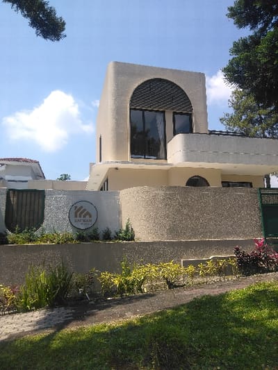 A modern, two-story building, likely residential or commercial, is depicted under a clear blue sky with scattered white clouds. The structure features a light beige and textured grey facade. The upper story presents a prominent rounded corner with a large, dark-framed arched window, detailed with an arched slatted screen above the glass panes. A curved balcony or terrace extends from the first floor. Below, textured retaining walls support the elevated property. A circular sign, bearing a black abstract logo and the text "KAIKAM HOUSE," is affixed to a lighter wall section. The foreground consists of a grassy slope, a dark paved path, and various low-lying plants, including long green blades, yellow-leafed shrubs, and reddish foliage. The scene is illuminated by sunlight, casting discernible shadows. Dark green tree foliage is visible in the upper corners of the image. No individuals are present. The location is Nová Bystřice, Czechia.