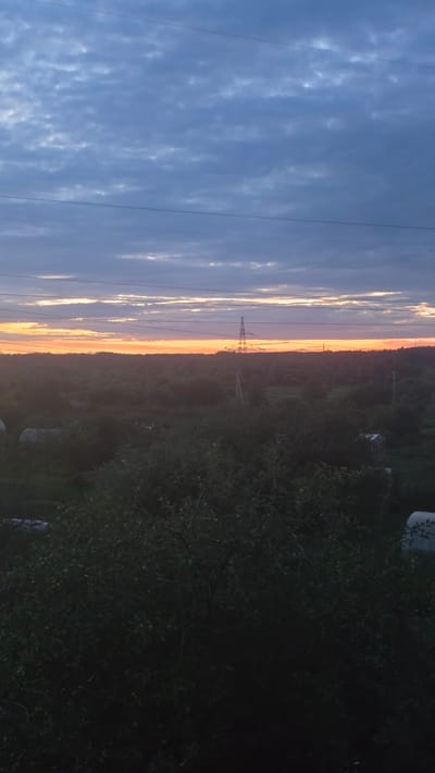 A vertical, eye-level, wide shot captures a dusky landscape in Yuryev-Polsky, Russia. The sky, a gradient of deep blue to pale orange, is filled with textured clouds. Power lines crisscross the upper portion of the frame. In the middle ground, a solitary power tower stands silhouetted against the horizon. The lower half of the image is dominated by dark foliage. The overall impression is one of twilight in a rural or semi-rural setting.