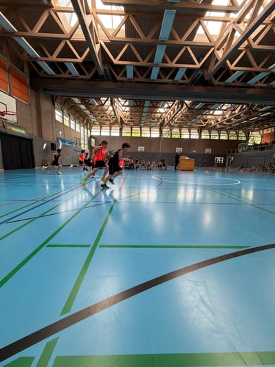 The image depicts the interior of a gymnasium located in Isny im Allgäu, Germany. The camera's viewpoint is from a low angle, pointing upwards at a slight oblique angle.

A group of young people, likely basketball players, are engaged in drills or exercises on the gymnasium floor. Some are wearing basketball gear, specifically jerseys.

The gymnasium floor is light blue with green and black lines delineating the court. The facility features a distinctive ceiling structure consisting of wooden beams. The walls have windows allowing natural light into the space. A wooden box is near one side of the court, potentially used for drills. There are also benches along the side, where other people can be seated, including a group wearing green.

The scene appears to be a basketball practice or training session.