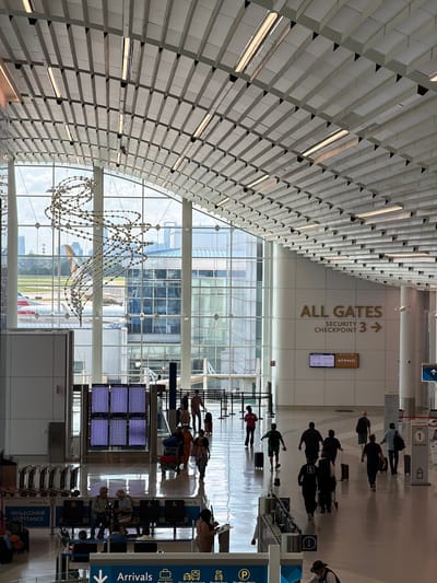 The image depicts the interior of a modern airport terminal in Charlotte, United States of America. The vast space is characterized by a high, vaulted ceiling with a complex, repeating geometric design and integrated lighting. Large windows offer a view of an airfield with airplanes visible.

In the foreground, the floor is highly reflective, mirroring the architectural elements above and the people below. Several departure boards displaying flight information are prominently positioned. A significant number of people are present, navigating the terminal. Some are walking with luggage, while others are seated on benches. There are also various airport amenities visible, including information kiosks, check-in counters, and baggage trolleys.

The environment suggests a bustling atmosphere, typical of an international airport. The overall aesthetic is clean and contemporary, with a focus on maximizing natural light through the extensive use of glass. The signage indicates directions for "ALL GATES," "SECURITY CHECKPOINT 3," and "DEPARTURES." The presence of "American" branding on seating and a sign suggests this may be within an American Airlines section of the terminal. The architectural style and overall ambiance are consistent with a large, well-equipped airport facility.