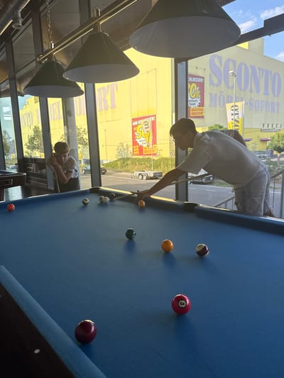 This image captures a casual moment at a pool hall, likely in Hanau, Germany, given the visible German text. The scene features two young men engaged in a game of pool. One person, wearing glasses and a light-colored shirt, is actively playing, leaning over the blue felt table with a cue stick in hand, preparing to strike a ball. The other individual, also young, is resting on the edge of the pool table, appearing relaxed and perhaps observing the game.

The setting is illuminated by natural light streaming through a large window, suggesting it is daytime, possibly late afternoon given the warm quality of the light. Through the window, a bright yellow building with large text is visible. The text on the building includes "SCONTO MÖBEL" and phrases like "WIR SCHLIESSEN!" (We are closing!), "10% VERKAUFS-RABATT" (10% sales discount), and "DRASTISCH REDUZIERT" (Drastically reduced), indicating a furniture store undergoing a sale. This commercial backdrop contrasts with the leisurely activity inside the pool hall.

Several billiard balls are scattered across the table, including a striped ball, a solid orange ball, and a red ball with the number '3'. The overall mood appears laid-back and social, characteristic of a friendly game among friends. The presence of modern cone-shaped lamps hanging from the ceiling adds to the contemporary atmosphere of the establishment.