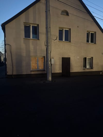 The image shows the exterior of a two-story building in Wąwelnica, Poland. The building has a light-colored facade and a dark sloped roof. There are several windows on the upper floor, and one window and a dark wooden door on the ground floor. The windows have white frames. The ground floor window on the left appears to have blinds, and the window on the right has blinds that are partially open. A utility pole with wires runs vertically along the right side of the building's facade. The area in front of the building is dark, suggesting it is nighttime or dusk, with a dark asphalt surface. A faint house number "17" is visible on the facade to the right of the door. The sky is a deep blue, indicating twilight.