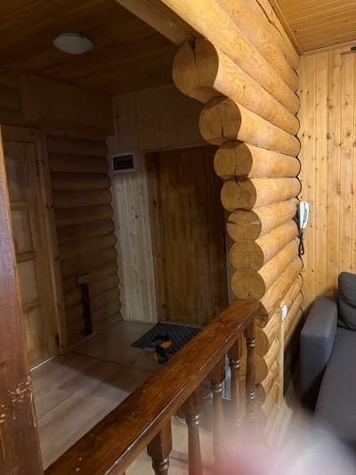 The image depicts an interior scene of a log cabin, likely in Novy Gorodok, Russia. The foreground features a dark wooden banister with visible wear and tear. Beyond the banister, a hallway is visible with wooden doors and walls constructed of stacked logs. A light-colored ceiling with a simple light fixture is overhead. To the right, a section of a dark gray couch is visible. On the wall of stacked logs, a white landline telephone with a coiled cord is mounted. On the floor in the hallway, a dark doormat with a pair of shoes on it can be seen. The overall aesthetic suggests a rustic and cozy interior.