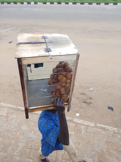A person carries a wooden and glass display case filled with golden-brown fried dough balls. The case has hinges on top and is open at the front, revealing the pastry-filled interior. A hand reaches into the case, holding a fork. The person is wearing a blue garment with a yellow swirl pattern and light-colored trousers. The scene appears to be outdoors, with a dusty dirt ground and a paved sidewalk in the foreground. In the background, a road with distinct black and white stripes along its edge is visible, suggesting a roadside location. The lighting suggests it could be daytime, and the weather appears to be fair. The overall impression is of a street vendor, likely in a marketplace or along a busy route in Nigeria, given the location context provided.